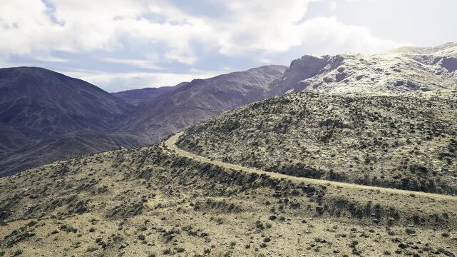 Winding namibia road leading to distant peaks, meandering trail hugging slope with stone scatter and layered ridges under broad cloud bank, sense of remote