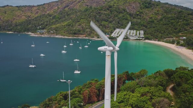 Wind turbine spinning near Windmill Viewpoint, lush green hillside overlooking tranquil bay, sailboats anchored in turquoise water and sandy shore leading to coastal resorts on Phuket island, Thailand
