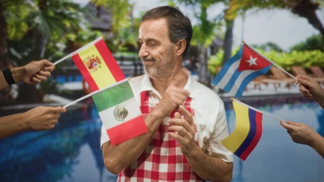Man in apron points finger to flag while hands hold small spain mexico cuba colombia flags in studio; identity indecision.