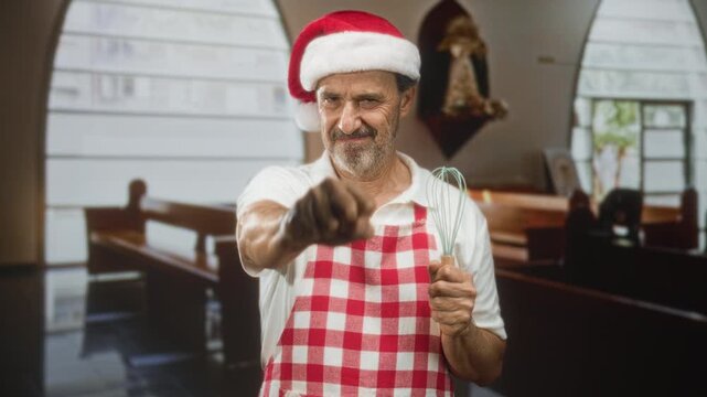 Man baker in santa hat and red checked apron whisking with hand while pointing a whisk toward camera in a building with pews and a religious statue; cheerful holiday.