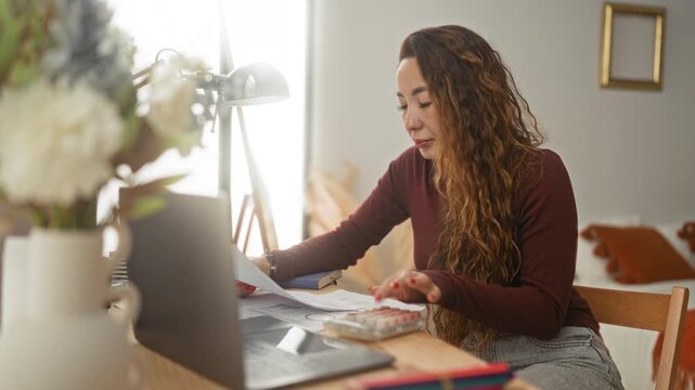 Woman at laptop holds printed pages and points finger to a calculator on a desk in a bedroom of a building with soft lamp light; concentration productivity.