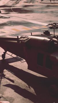 A military helicopter is parked on a city street. The helicopter has a sleek design and is surrounded by shadows from nearby buildings. People walk by unaware of the aircraft.