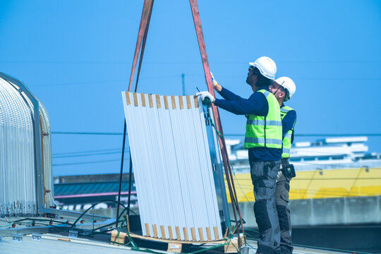 A renewable energy specialist in reflective workwear and helmet confidently unhooks the heavy lifting rigging from the panel bundle on a large corrugated factory rooftop under the tropical.