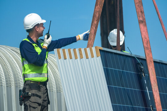 Two renewable energy specialists in hard hats and hi vis workwear precisely direct the crane over the module stack by radio high on an industrial warehouse roof in a Thai city.
