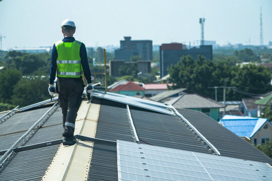 A photovoltaic technician wearing a safety vest and hard hat steadily pauses at the roof edge platform between inspections on the curved metal roof of an industrial factory in Bangkok.