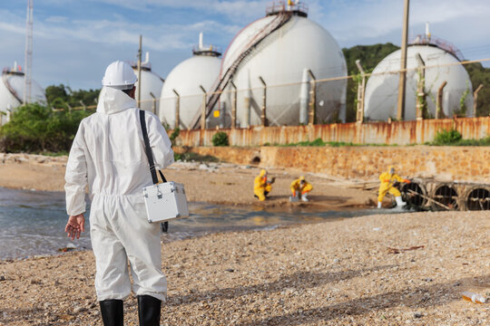 Environmental inspector wearing protective clothing and carrying field equipment while supervising a team conducting a coastal environmental survey near an industrial storage facility.