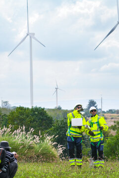 Two wind farm surveyors with laptop and rolled drawings check micro siting positions against the plan in tall grass on a ridge inside the wind farm in bright tropical daylight at a wind.