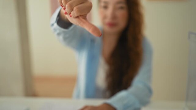 Woman shows thumbs down gesture with visible thumb and red nails at a table in a building; disapproval.