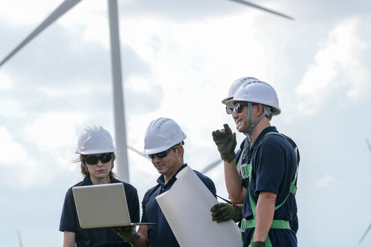 A four person rigging crew in navy suits and white helmets debate the lift plan while carrying the load under turning rotors on the wind farm in bright hazy daylight at an operating.