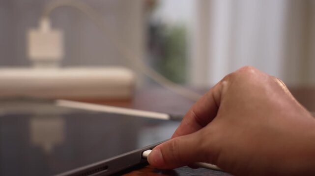 Close up shot of digital tablet on wooden desk as hand plugs charging cable, dark display screen turning green with power adapter blurred in home workspace background, smart home technology concept.