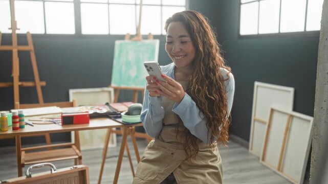 Woman wearing apron holds smartphone and pumps fist in artist studio with easels and paint bottles; creative joy.