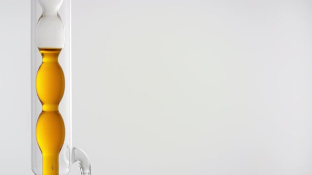 Close shot of transparent orange liquid drains off a vertically mounted bulb laboratory condenser on the left side of the frame, leaving the right side as copy space on grey background
