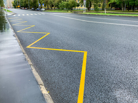 A freshly renewed yellow zigzag marks the bus stop zone along the kerb of a wet city street. New asphalt and white lane lines shine after the rain. Urban road infrastructure