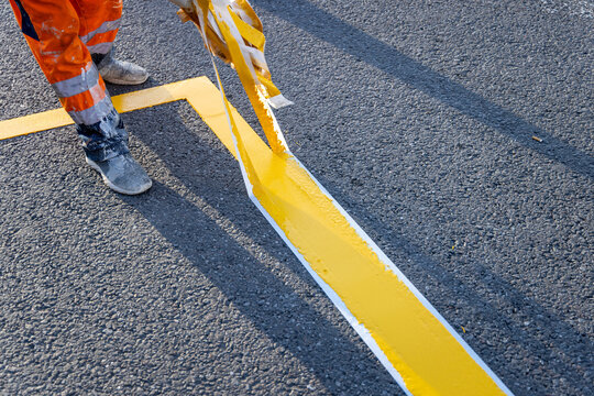 A road worker in an orange high visibility suit is pulling masking tape off the fresh yellow line. The bright marking stands out on the grey asphalt. Everyday road maintenance work