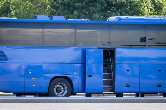 A modern intercity coach waits at the kerb with its passenger door open. The long blue body reflects the sunlit trees behind. Tourist transfers, charter trips and road travel by bus
