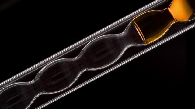 Close-up shot of orange transparent liquid inside of the inclined bulb laboratory condenser over black backdrop | Propolis tincture cosmetic formulation concept