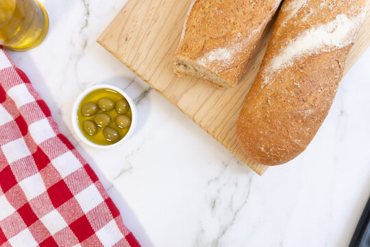 Fresh bread rests on a wooden board. Small bowl holds green olives in olive oil. Red and white checkered cloth adds charm. White marble background enhances simplicity