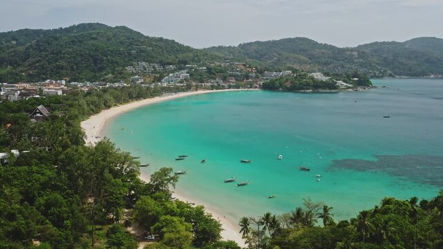 Kata Beach features a long white sand coastline with turquoise ocean water, longtail boats floating, and a lush green mountain landscape enclosing the tropical bay in Thailand. Aerial drone flight