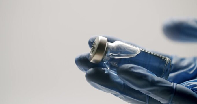 Healthcare worker's hand in a blue latex glove holding a small glass vial with transparent medicine, preparing for vaccination on a clean, isolated white background laboratory setting