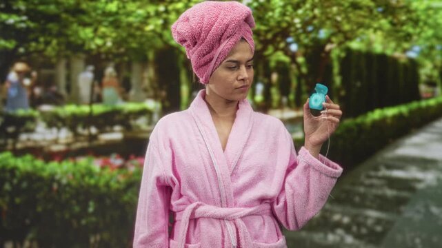 Woman in pink bathrobe with towel turban holds dental floss container in hand on tree lined street; calm self care.