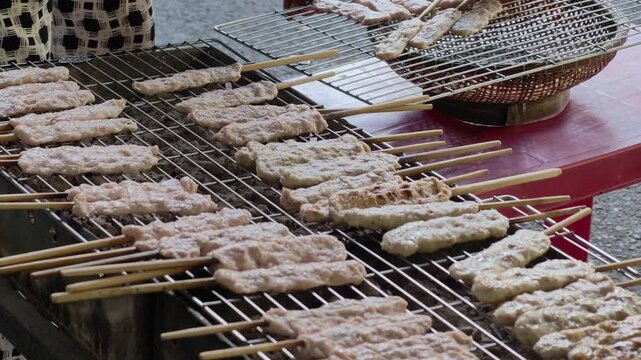 Medium shot of multiple rows of Vietnamese pork skewers nem nuong being grilled on a large charcoal wire mesh grill at a busy street food stall