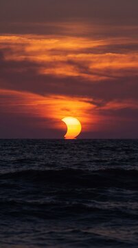 Partial solar eclipse at sunset over a dark ocean horizon during a vibrant golden hour twilight sky