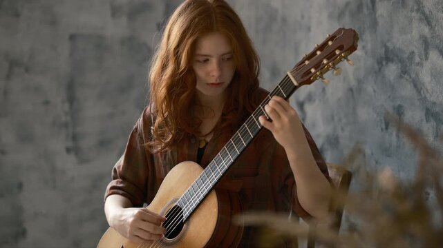 Cinematic portrait of a young redhead girl playing an classic guitar. She wears a casual plaid shirt and focuses intently on the strings against a grey textured wall. The shot features soft lighting 
