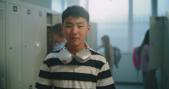 Portrait of Friendly Asian Teenage Boy Student with Wireless Headphones Waving Hand and Looking at Camera in School Corridor. Campus Atmosphere, Responsibility, Familiar Routines of Teenage Education.