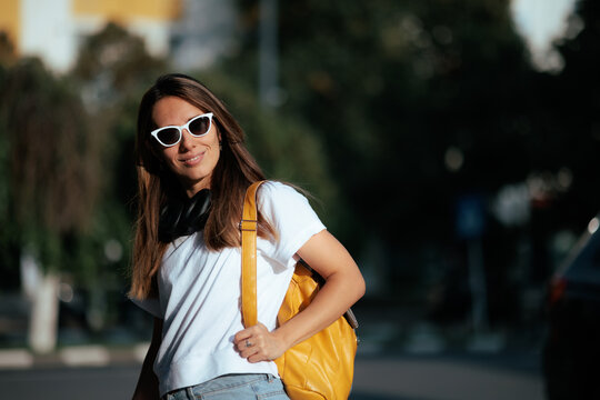 Smiling Young Woman Wearing White Sunglasses And Holding Yellow Backpack. Attractive cheerful girl carrying trendy leather bag while walking outside on sunny day 