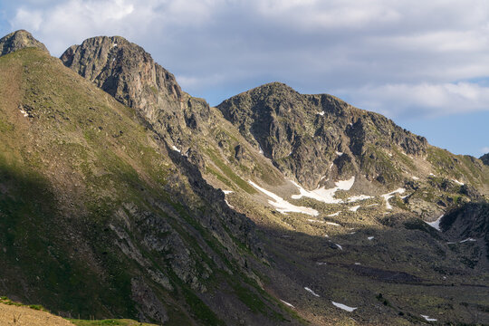 Craggy Pyrenean ridges surround a rugged glacial valley in Andorra filled with scattered rocks and lingering snow patches. Sunlit slopes and dramatic rocky peaks stretch beneath a cloudy summer sky
