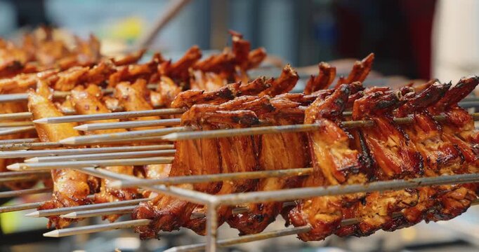 Grilled chicken wings on metal skewers at an outdoor street food market. Close-up of roasted poultry meat, traditional Asian cuisine, barbecue cooking process, commercial food background.