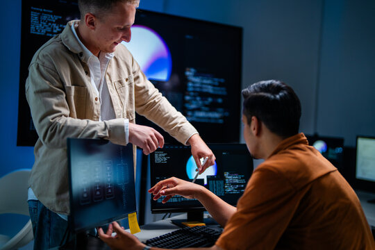 Two colleagues in a security operations centre discuss a finding while facing the display at a multi-monitor workstation in a darkened control room, defending the corporate network.