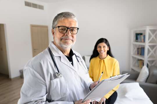 Senior male doctor in uniform holds clipboard during consultation in clinic office, provides medical services and treatment plan to female patient, posing for portrait at hospital. Healthcare concept