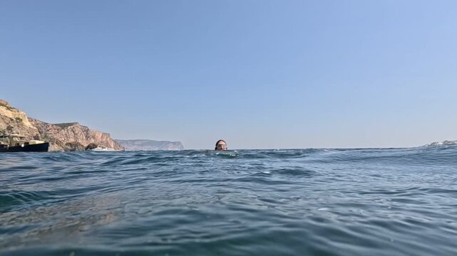 Person swimming in choppy sea near dramatic rocky cliffs under clear blue sky. Low angle shot.