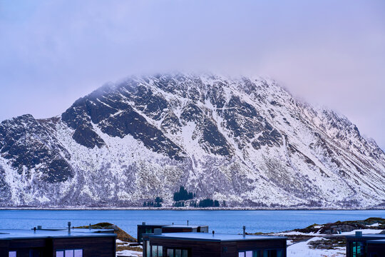 Majestic winter mountain peak glowing in spectacular golden hour light in Lofoten, Norway. Beautiful scenic view of rugged snow-covered Arctic mountains illuminated by warm sunrise or sunset under a d
