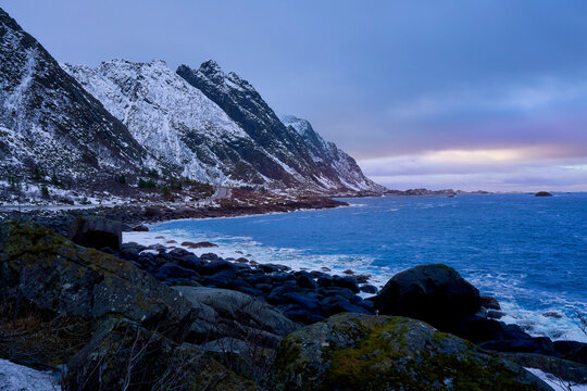 Majestic winter landscape of Lofoten islands in Norway with bright sunlight reflecting on the calm blue sea. Beautiful scenic view of grand snow-covered mountains under a bright dramatic sky in Arctic