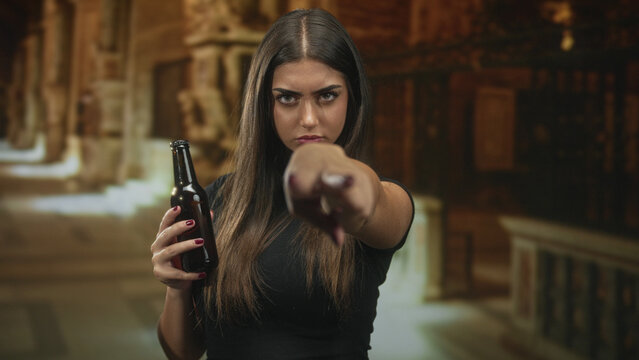 Woman holding beer bottle and making v sign over eyes while pointing finger toward camera in building; defiance confrontation.