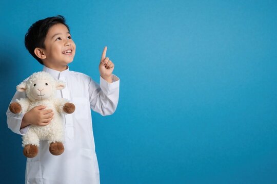 Asian Muslim boy wears white thobe holding sheep plush toy. Smiling child points up on blue background, ready for Eid Al Adha celebration. This image is perfect for festive greetings.