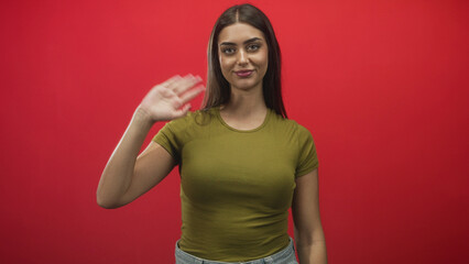 Woman waves right hand in studio against red backdrop wearing green t shirt and jeans, smiling and making a clear greeting gesture  welcoming. © Krakenimages.com