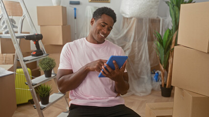 Man tapping a tablet with finger while smiling, seated among packed boxes, ladder and drill inside a building  moving day contentment. © Krakenimages.com