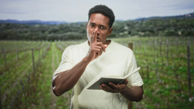 Man holds an open book and presses finger to lips for silence in a vineyard forest near rolling hills; quiet contemplation.