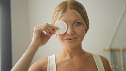 Woman holding cotton pad over eye in studio, smiling with exposed neckline and hand raised in skincare gesture  self care calm. © Krakenimages.com