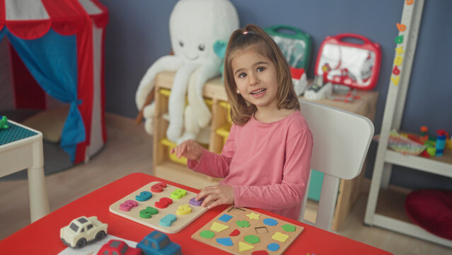 Girl uses right hand to place colorful wooden puzzle piece on peg board at a red table in a building; concentration learning play.
