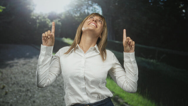 Woman points index fingers upward on forest path, smiling with sunlit hair and a white shirt; optimism discovery.