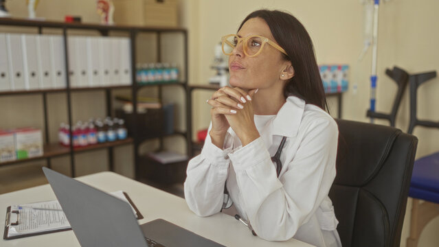 Woman doctor with stethoscope touches chin with finger while seated at laptop in building; thoughtful reflection.