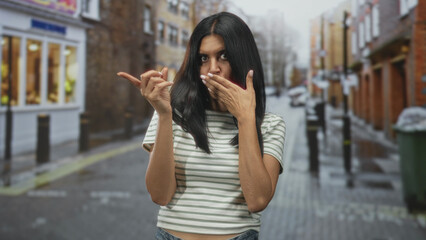 Fototapeta premium Woman with hand covering mouth and other hand making a beckon gesture on a wet cobbled street; playful flirtation.
