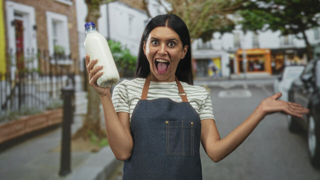 Woman in apron holding a milk bottle in hand, making a scrunched face and raising the bottle toward camera on a busy street with parked cars; playful defiance.