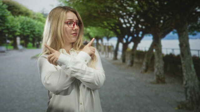 Woman pointing crossed index fingers on a tree lined street by the waterfront; uncertainty hesitation.
