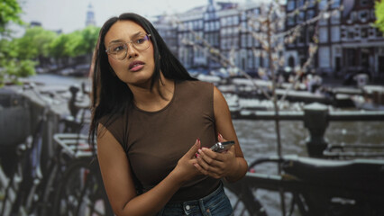 Fototapeta premium Woman brunette wearing glasses holding smartphone with visible hands and painted nails, glancing aside while seated in a studio with canal backdrop; pensive.