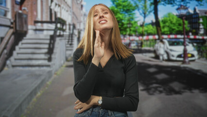 Fototapeta premium Woman clasping hands to mouth on a sunlit city street with bare midriff visible and watch on wrist, distressed expression and closed eyes; despair pleading.